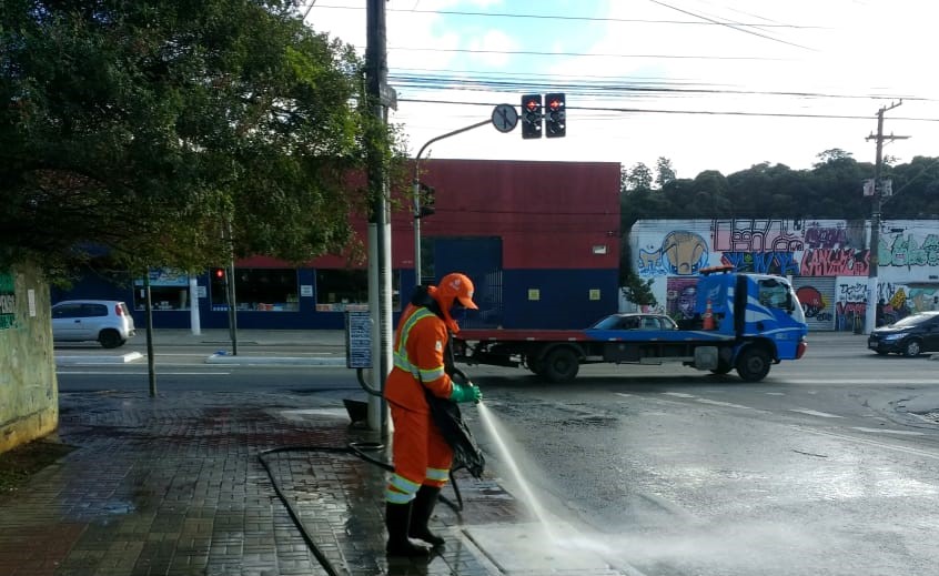 Homem, com roupas e equipamentos de proteção, segura mangueira com jato d'água, lavando calçada. À frente uma carreta está passando na rua.